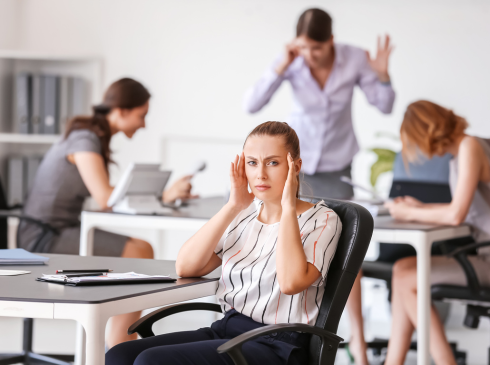 Inside an office. A woman in the foreground hold her hands to her temples as if she has a headache. In th ebackground out of focus are busy employees.
