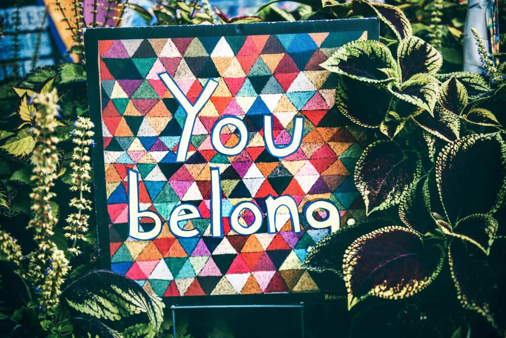 Sign reads "you belong". Multicoloured background Plants surround the sign.