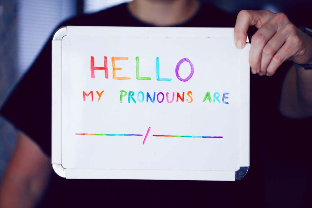 Someone holds a sign that read 'hello my pronouns are'. The letters on the whiteboard and rainbow coloured.